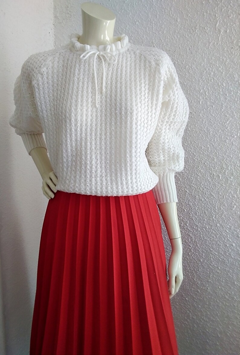 May include: A white knitted sweater with a ruffled neckline and a tie at the neck, paired with a red pleated skirt.