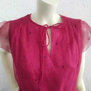May include: A magenta blouse with a keyhole neckline and tie detail. The top has short, sheer, ruffled sleeves and is decorated with small, dark purple polka dots. The garment is displayed on a mannequin.