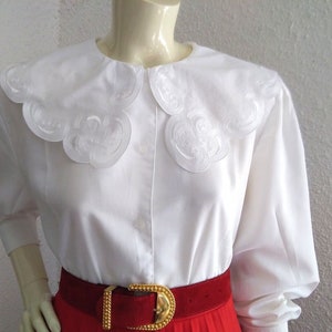Puede incluir: Blusa blanca de manga larga con un cuello grande blanco festoneado con detalles bordados. La blusa se lleva con un cintur&oacute;n rojo.