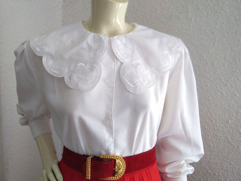 Puede incluir: Blusa blanca de manga larga con un cuello grande blanco festoneado con detalles bordados. La blusa se lleva con un cintur&oacute;n rojo.