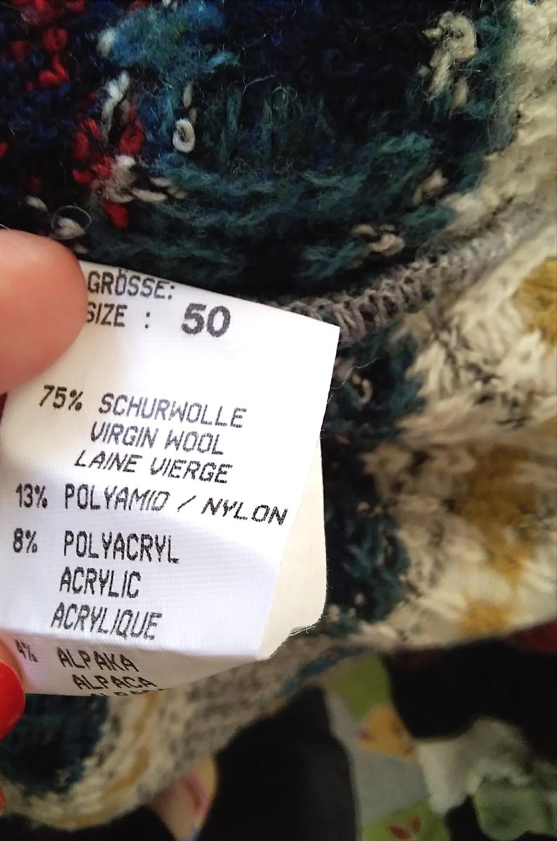 May include: A white tag with black text detailing the composition of a sweater. The tag states "GR&Ouml;SSE: SIZE: 50" and lists the percentages of Schurwolle (Virgin Wool), Polyamid (Nylon), Polyacryl (Acrylic), and Alpaka (Alpaca).