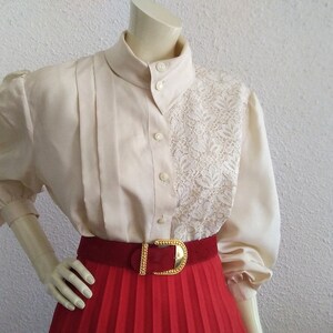 May include: A white blouse with a lace panel and a red pleated skirt. The blouse has a high collar and long sleeves with puffed shoulders. The skirt has a gold buckle belt.