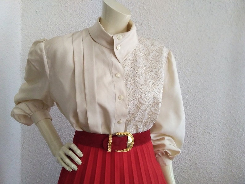 May include: A white blouse with a lace panel and a red pleated skirt. The blouse has a high collar and long sleeves with puffed shoulders. The skirt has a gold buckle belt.