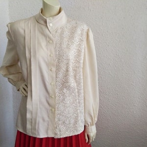 May include: A cream-colored blouse with a lace panel on the right side. The blouse has a high collar and long sleeves.