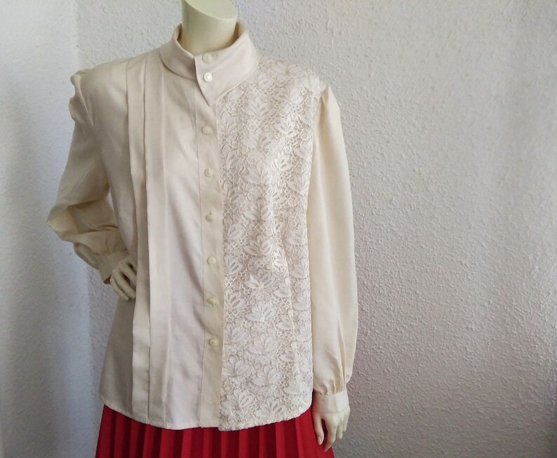 May include: A cream-colored blouse with a lace panel on the right side. The blouse has a high collar and long sleeves.