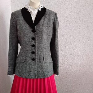 May include: A grey tweed blazer with a black velvet collar and black buttons. The blazer is paired with a bright pink pleated skirt and a white lace-trimmed blouse. The outfit is displayed on a mannequin.