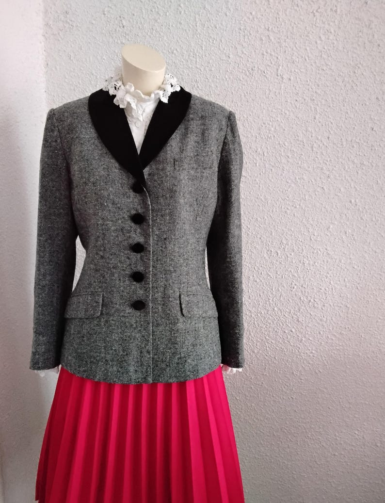 May include: A grey tweed blazer with a black velvet collar and black buttons. The blazer is paired with a bright pink pleated skirt and a white lace-trimmed blouse. The outfit is displayed on a mannequin.