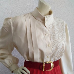 May include: A white blouse with a lace panel on the right side. The blouse has a high collar and long sleeves with puffed shoulders. The blouse is buttoned up the front.
