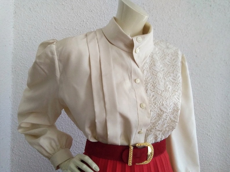 May include: A white blouse with a lace panel on the right side. The blouse has a high collar and long sleeves with puffed shoulders. The blouse is buttoned up the front.