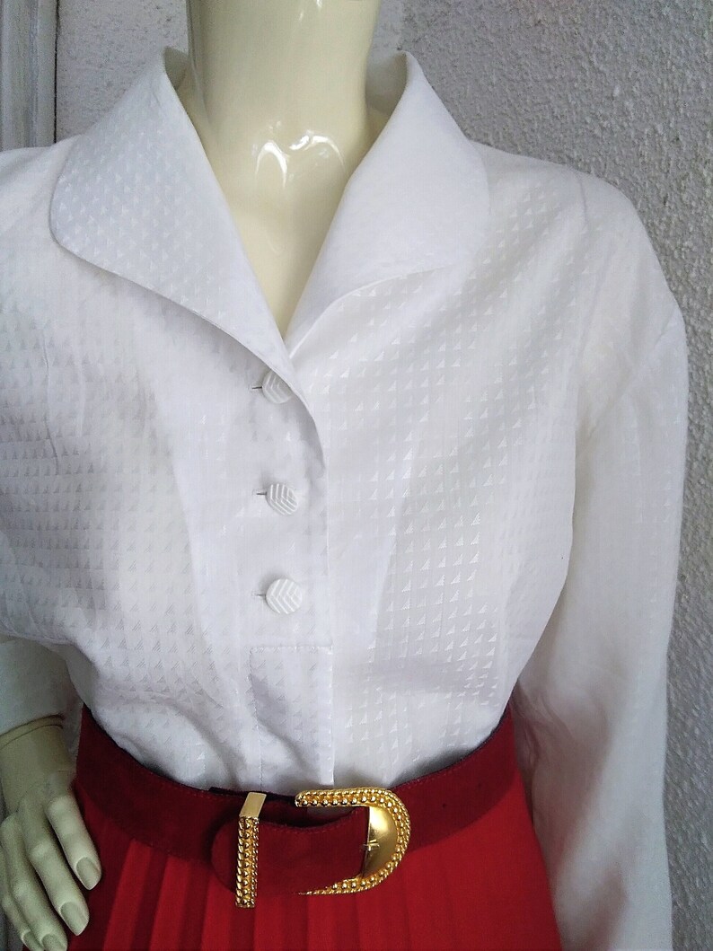 May include: White button-down blouse with a textured pattern and a wide collar. The blouse is worn with a red skirt and a gold belt.