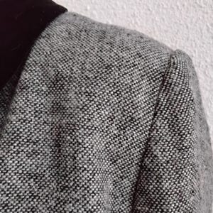 May include: A close-up shot of a grey tweed blazer featuring a black velvet collar. The blazer has a textured, speckled pattern in shades of grey and black. The shoulder seam is visible, suggesting a tailored fit. The garment appears to be made of a woven fabric.