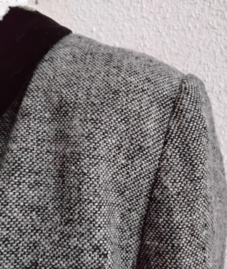 May include: A close-up shot of a grey tweed blazer featuring a black velvet collar. The blazer has a textured, speckled pattern in shades of grey and black. The shoulder seam is visible, suggesting a tailored fit. The garment appears to be made of a woven fabric.