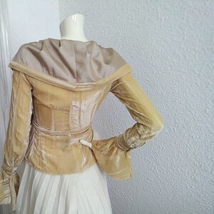 May include: A gold velvet jacket with a wide, off-the-shoulder collar and long sleeves. The jacket has a fitted waist and a slightly flared hem. The sleeves have a gathered detail at the cuffs.
