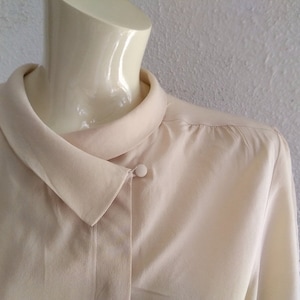 May include: A cream-coloured, long-sleeved blouse with a button-up collar and a single button closure at the neck.