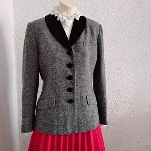 May include: A grey tweed jacket with a black velvet collar and black buttons. The jacket has two front pockets and is worn with a bright pink pleated skirt. A white lace collar is visible beneath the jacket.