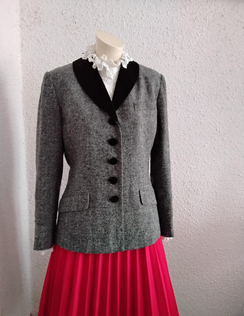 May include: A grey tweed jacket with a black velvet collar and black buttons. The jacket has two front pockets and is worn with a bright pink pleated skirt. A white lace collar is visible beneath the jacket.