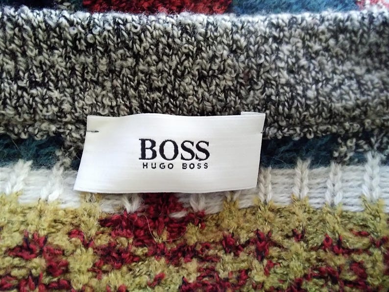 May include: A close-up of a white label with black text that reads "BOSS HUGO BOSS" on a gray and white knit sweater with a colorful pattern.