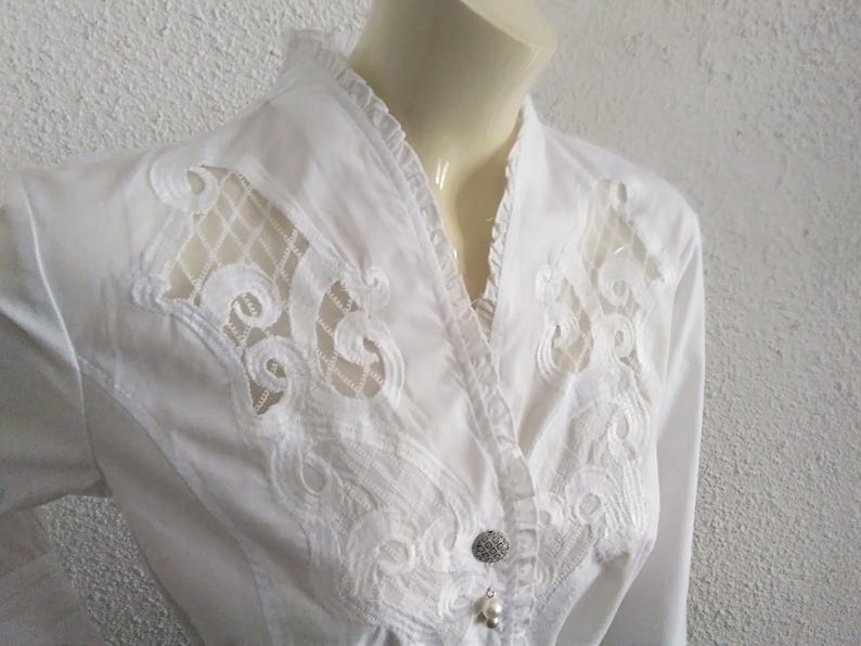 K&ouml;nnte beinhalten: Wei&szlig;e Bluse mit einer Spitzen&uuml;berlagerung und einem Perlenknopf-Detail.