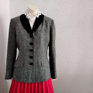 May include: A grey tweed blazer with a black velvet collar and four black buttons. The blazer has two front pockets and is displayed on a mannequin. A white blouse with a lace collar is visible beneath the blazer. A red pleated skirt is visible at the bottom.