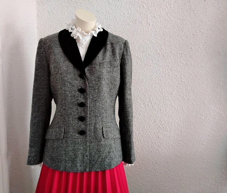 May include: A grey tweed blazer with a black velvet collar and four black buttons. The blazer has two front pockets and is displayed on a mannequin. A white blouse with a lace collar is visible beneath the blazer. A red pleated skirt is visible at the bottom.