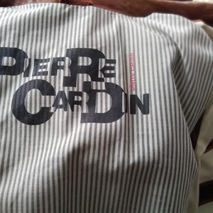May include: A white and gray striped fabric with the text "PIERRE CARDIN" printed in black. The fabric has a red and white striped tag with the text "pierre cardin.com" printed in black.