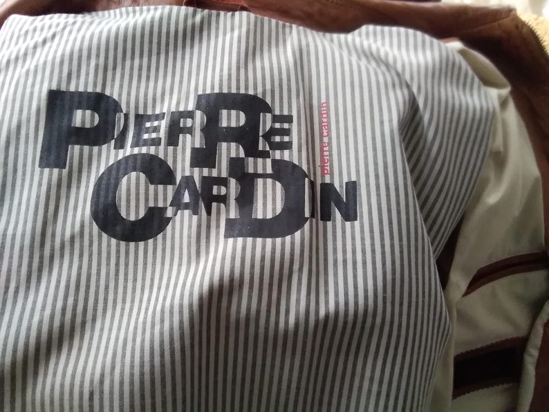 May include: A white and gray striped fabric with the text "PIERRE CARDIN" printed in black. The fabric has a red and white striped tag with the text "pierre cardin.com" printed in black.