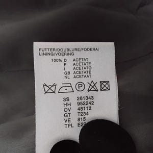 May include: A close-up of a white clothing label with black text. The label details washing instructions, fabric composition, and codes. The label reads "FUTTER/DOUBLURE/FODERA/LINING/VOERING" and "100% D ACETAT".