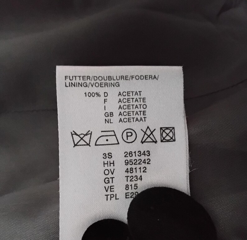 May include: A close-up of a white clothing label with black text. The label details washing instructions, fabric composition, and codes. The label reads "FUTTER/DOUBLURE/FODERA/LINING/VOERING" and "100% D ACETAT".