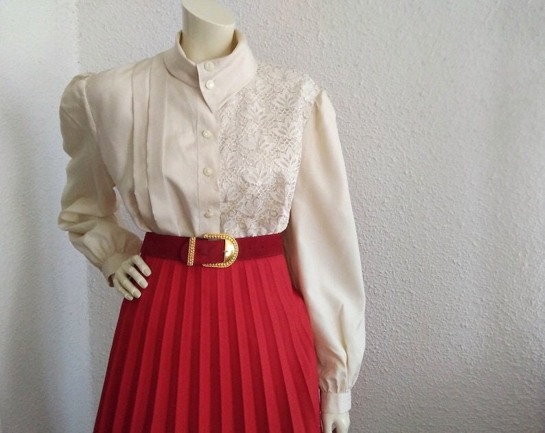 May include: A white blouse with a lace overlay and a red pleated skirt. The blouse has a high collar and long sleeves. The skirt has a gold belt.