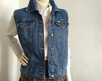 Op de afbeelding: Een blauwe denim vest met knoopsluiting en twee zakken. De vest wordt gedragen over een wit blouse met een kantpatroon.
