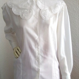 Puede incluir: Una blusa blanca de manga larga con un cuello grande, blanco y festoneado. El cuello tiene un delicado dise&ntilde;o bordado.