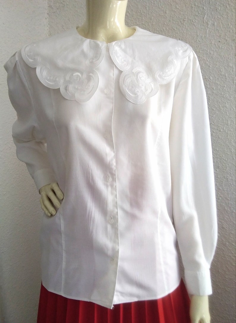 Puede incluir: Una blusa blanca de manga larga con un cuello grande, blanco y festoneado. El cuello tiene un delicado dise&ntilde;o bordado.