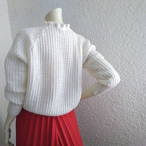 May include: White knitted sweater with a ruffled neckline and a red pleated skirt.