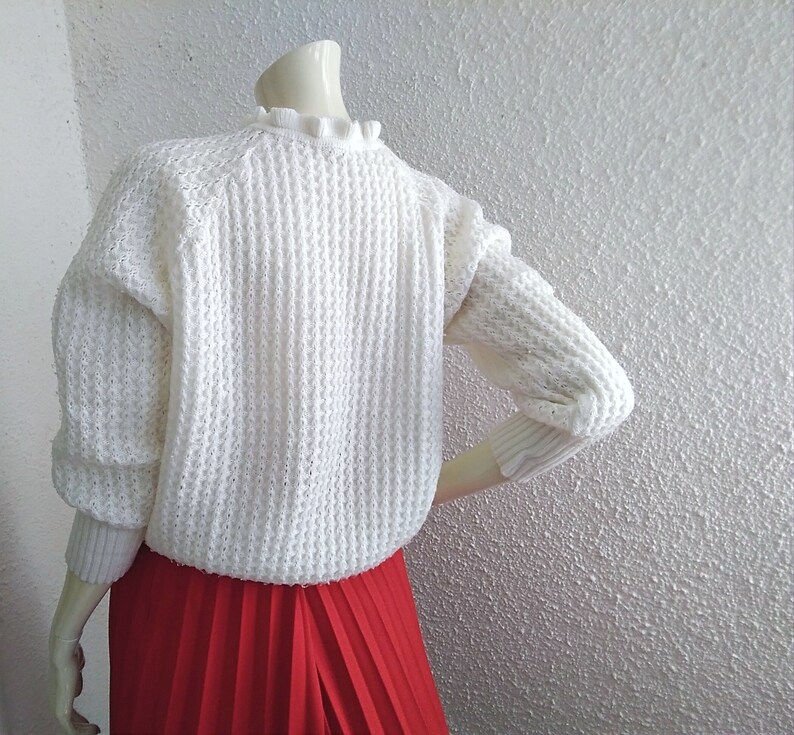 May include: White knitted sweater with a ruffled neckline and a red pleated skirt.