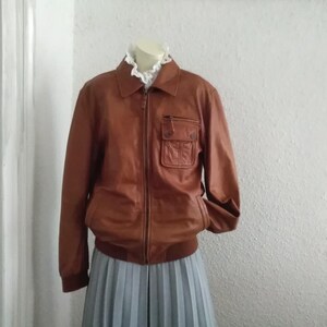 May include: A brown leather jacket with a zipper closure and a large pocket on the left side. The jacket has a stand-up collar and long sleeves.