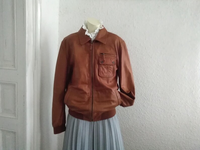 May include: A brown leather jacket with a zipper closure and a large pocket on the left side. The jacket has a stand-up collar and long sleeves.