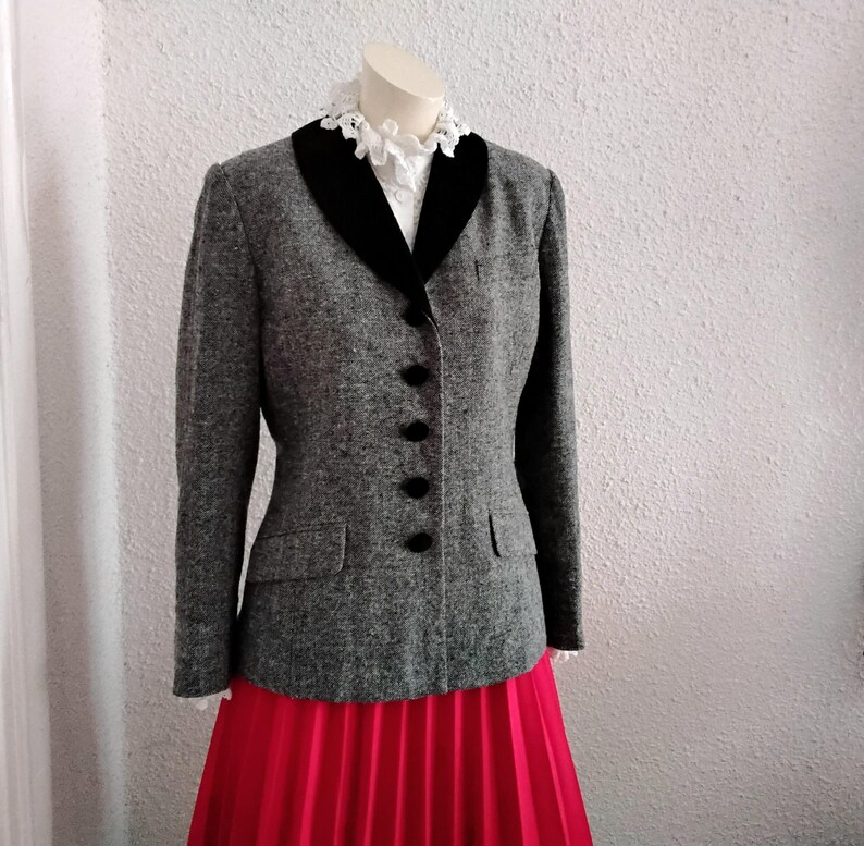 May include: A grey tweed jacket with a black velvet collar and black buttons. The jacket is displayed on a mannequin over a white blouse with a lace collar and a red pleated skirt. The jacket has two front pockets.