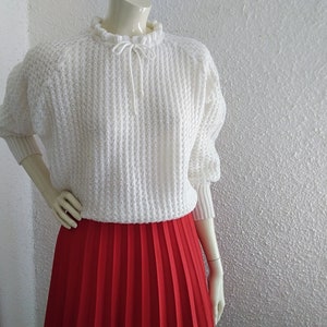May include: White knitted sweater with a ruffled neckline and a tie at the neck. The sweater has a loose fit and long sleeves. The model is wearing a red pleated skirt.