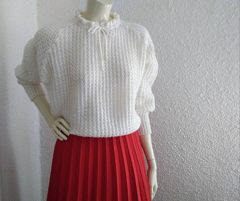 May include: White knitted sweater with a ruffled neckline and a tie at the neck. The sweater has a loose fit and long sleeves. The model is wearing a red pleated skirt.