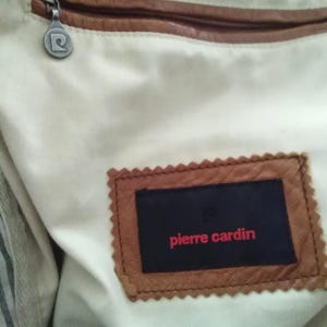 May include: A brown leather bag with a zipper closure and a white lining. The bag has a brown leather label with the text "pierre cardin" in red.