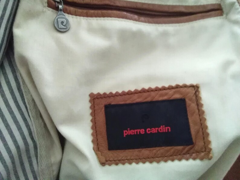 May include: A brown leather bag with a zipper closure and a white lining. The bag has a brown leather label with the text "pierre cardin" in red.