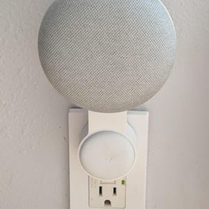 May include: A white smart speaker mounted on a white wall outlet. The speaker is a light gray color with a fabric-like texture. The outlet has a green light illuminated.