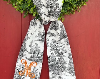 May include: A white and black toile fabric bow with an orange embroidered letter "M" in the centre. The bow is tied around a green wreath.