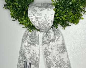 May include: A white fabric ribbon with a grey toile pattern and the letter "M" embroidered on it. The ribbon is tied around a green wreath.