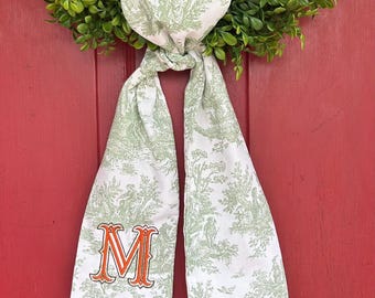 May include: A white fabric door wreath with a green toile pattern and an orange embroidered letter "M".