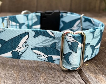 May include: A light blue dog collar with a white shark pattern. The collar has a black buckle and a gold metal D-ring.
