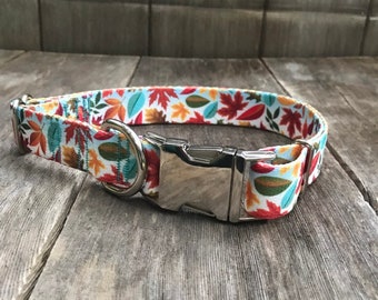 May include: A white dog collar with a colorful autumn leaf pattern. The collar has a silver buckle and a D-ring.