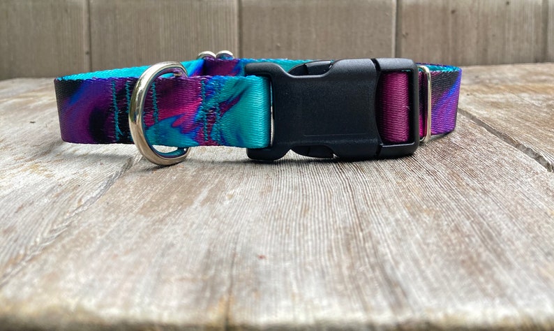 May include: A purple, blue, and black patterned dog collar with a silver D-ring and a black plastic buckle.