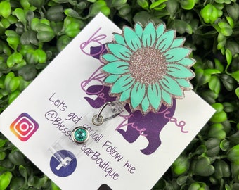 May include: A teal glitter sunflower badge reel with a green gem charm. The reel is attached to a white card with the text "Let's get social! Follow me @BlessWearBoutique" and social media icons.