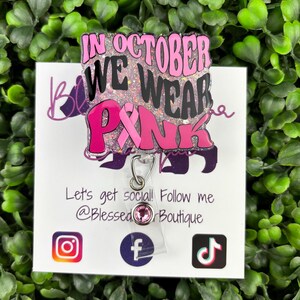 A flat lay image displays a badge reel with the words "IN OCTOBER WE WEAR PINK" in bold, black and pink lettering. A pink ribbon symbol is incorporated into the design. The background of the badge reel is a light pink glitter.